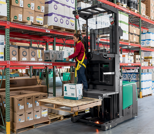 A woman in a red shirt operates a teal and gray forklift, reaching for boxes on a high shelf in a warehouse with metal shelving.