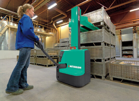 Indoor shot of a woman operating a teal Mitsubishi forklift in a large warehouse.